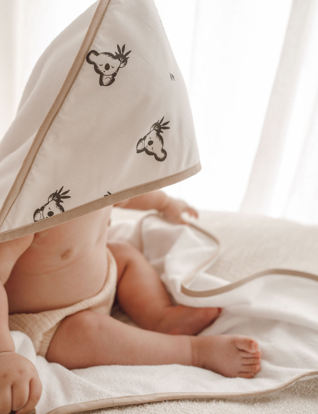 Bébé enveloppé dans une cape de bain blanche à motifs koala, allongé sur un linge blanc.