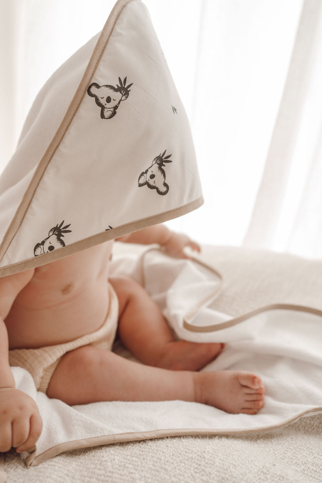 Bébé enveloppé dans une cape de bain blanche à motifs koala, allongé sur un linge blanc.